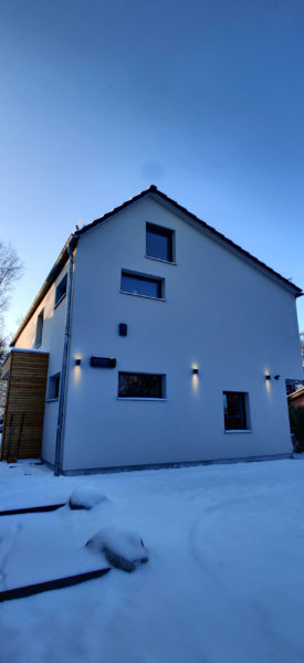 Moderne Hausfassade mit Wandbeleuchtung im Schnee neben einer hohen Hecke im Winter