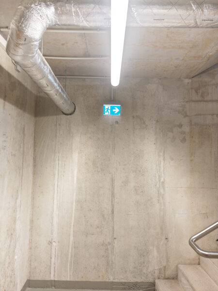 Treppenhaus der Wohnanlage Sheridan Park & Junia GbR mit Notausgangsschild, LED-Leuchte und Lüftungsrohren