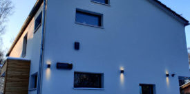 Moderne Hausfassade mit Wandbeleuchtung im Schnee neben einer hohen Hecke im Winter